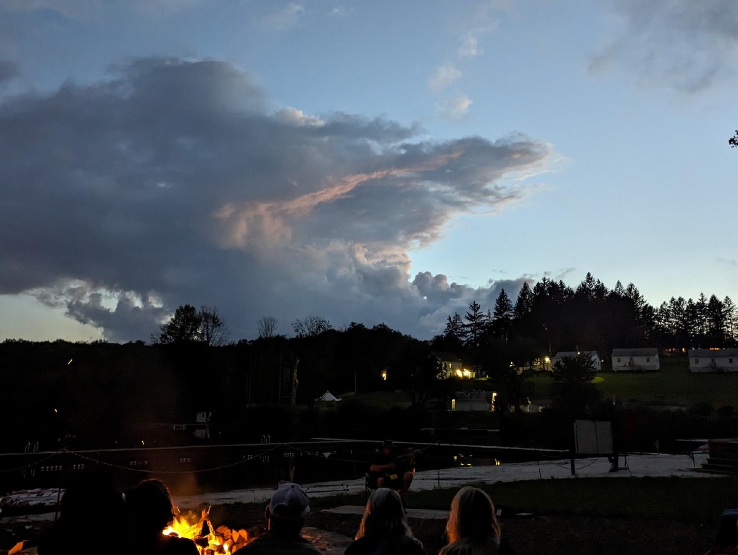 A man, seated, performs a song at sunset. In the foreground are the silhouettes of several people, facing away as they listen to the music. In the distance a hill rises away with several white buildings on it. An impressive cloud formation looms overhead.